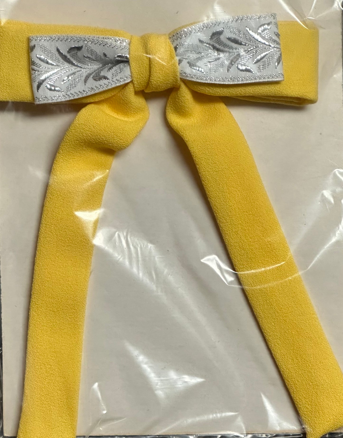 Experience the charm of the South with the Colonel Ties, crafted by Square Up Fashions. This bow tie showcases a sunny yellow bow with elegant long tails and a sophisticated silver patterned center, all beautifully displayed against a sleek plastic-wrapped background.