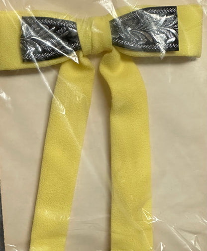 A Colonel Ties bow from Square Up Fashions, featuring a yellow and silver pattern and long tails reminiscent of a classic southern bow tie, is wrapped in clear plastic packaging and lies on a flat surface.